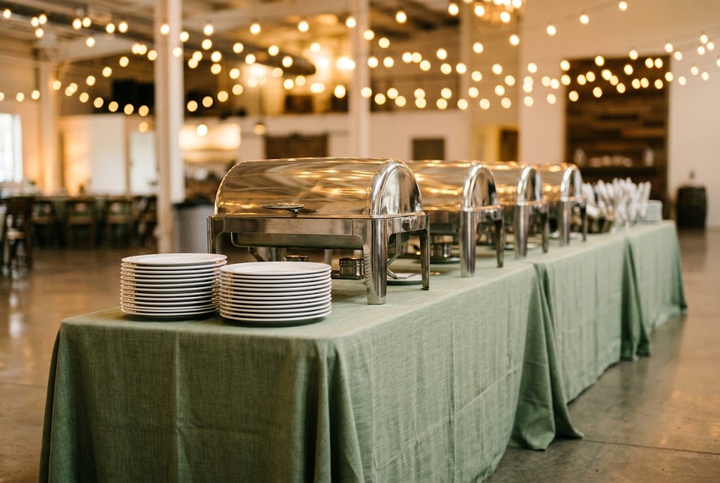 Banquet table buffet setup with roll-top chafing dishes and stacked white plates on sage green linen in a Bay Area event hall